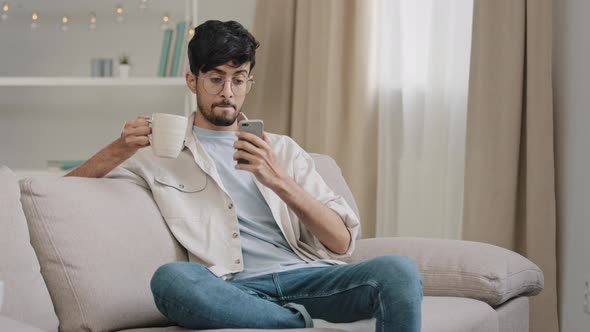 Millennial Arabic Guy Indian Handsome 30s Man Sitting at Home on Couch Drinking Hot Tea Coffee alt