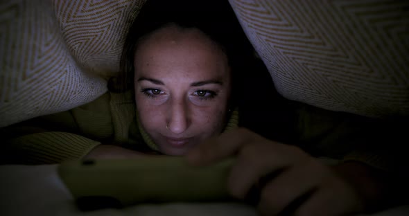 Woman on the bed watching movie on smartphone staying under the blanket