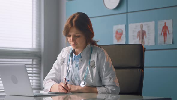 Brunette general practitioner in white medical coat writes in notebook alt
