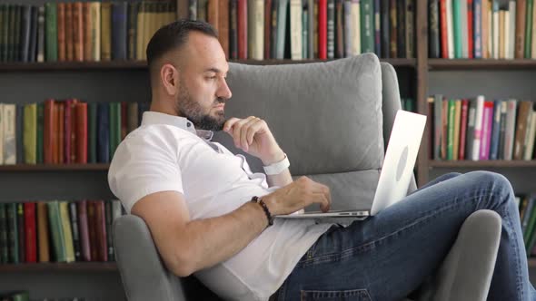 Mature Man Is Working on Laptop Sitting in Armchair at Home Office, Side View.