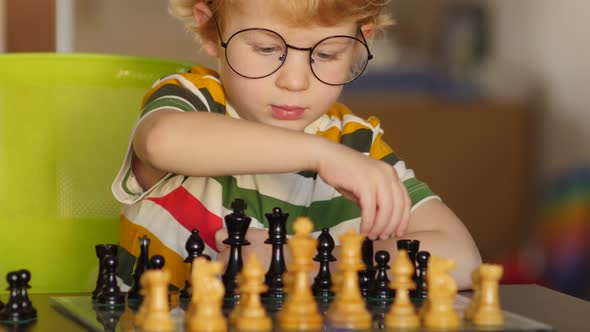 Funny young blonde pre-school boy with glasses plays chess. alt