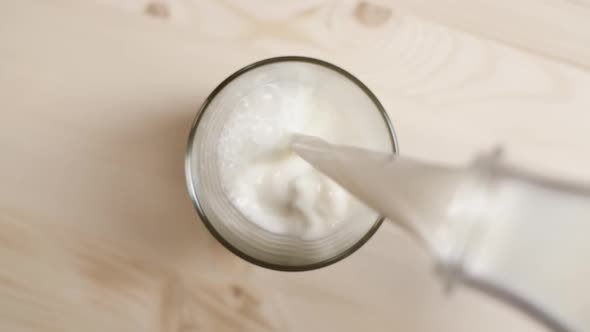 Top View of Milk Being Poured From Transparent Bottle in a Glass on Light Brown Wooden Table alt