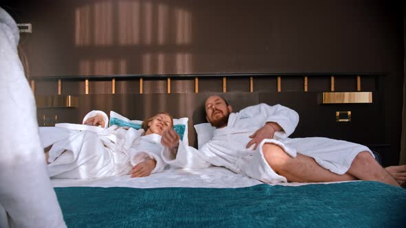 A Happy Successful Parents in White Bathrobes Falling on the Bed and Their Daughter Creeps Up To alt