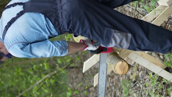 Vertical Shot Woodcutter in Working Uniform Sawing Tree Trunk on Sawhorse with Electric Saw