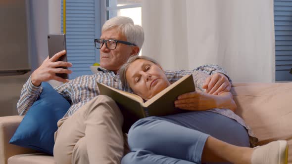 Happy Aged Married Couple Relaxing Together at Home with Book and Cellphone alt