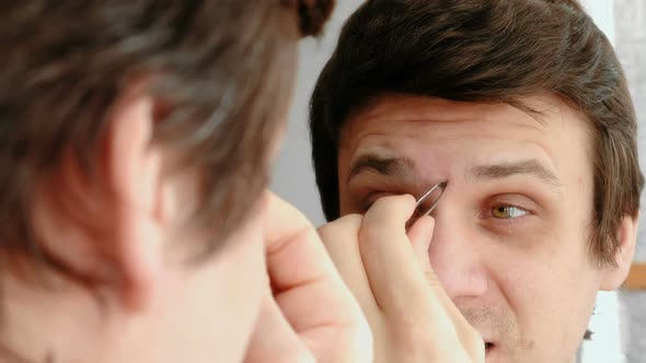 Young Man Plucking His Eyebrows with Tweezers and and Winces From the Pain. Styling Eyebrows.