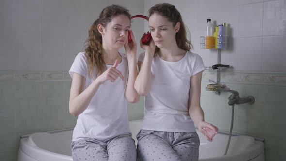 Front View Identical Twin Sisters Listening to Music in Headphones Sitting in Bathroom on Bathtub alt