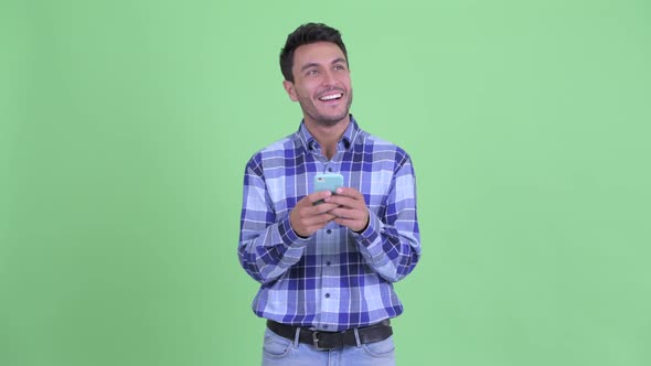 Happy Young Hispanic Man Thinking While Using Phone alt