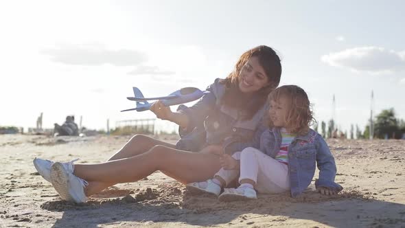 Mother and Daughter Sitting on the Beach and Play with Toy Plaine. Happy Family Concept