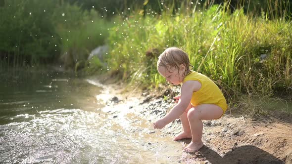 Little Funny Cute Blonde Girl Child Toddler in Yellow Wet Bodysuit Playing By the Lake Waterside