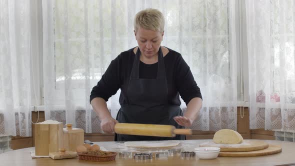Middle-aged Woman Rolls Dough with Wooden Rolling Pin on Flour Sprinkled Table alt