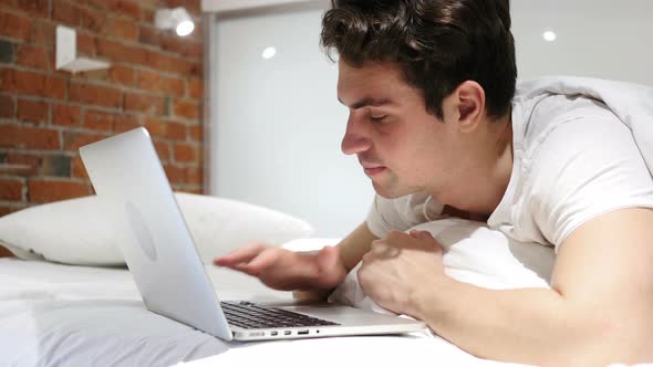 Man in Bed Working on Laptop and Reacting to Loss, Failure alt