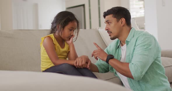 Hispanic father telling off his young upset daughter sitting on sofa alt