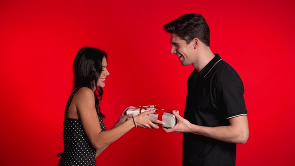 Young Couple Exchange Gifts on Red Studio Background. Love, Holidays Concept. alt
