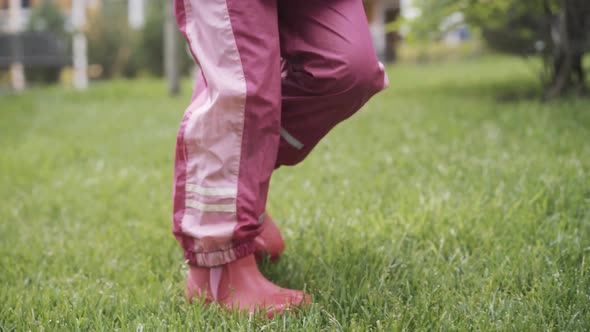 Feet of Little Girl in Pink Rubber Boots and Waterproof Pants Walking on Wet Green Grass Outdoors alt