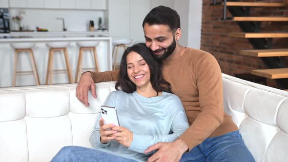 Indian Couple Sitting on the Sofa in Cozy Living Room Using Phone for Virtual Connectio