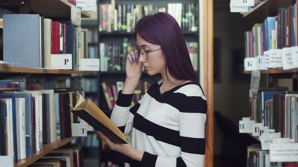 Female student with dark hair adjusts her glasses and reads a book, standing in the library, between alt