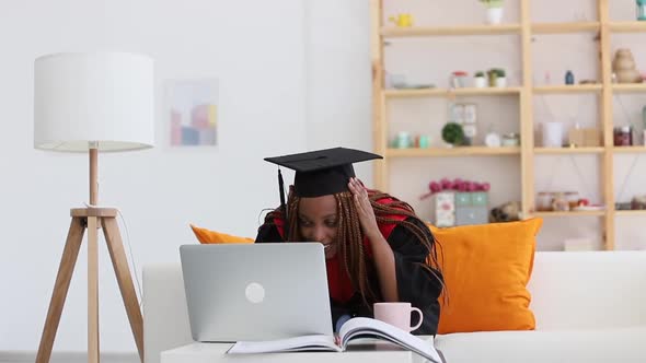 Female Student Wearing Cap and Gown Has Online Talk at Table with Computer at Home Room Spbi alt