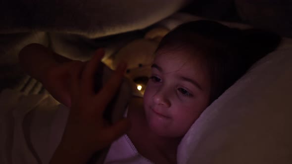 Little girl lies in bed at night and plays games on the phone.
