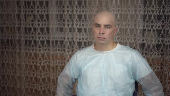 A Bald Young Man with Oncology Looks Sadly at the Camera. Man in a Wheelchair at Home. Hair Loss Due alt