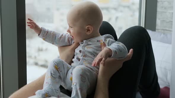 father plays with baby son sitting on his stomach by the window