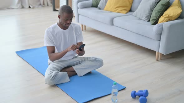 Young African Man Using Smartphone on Yoga Mat at Home alt
