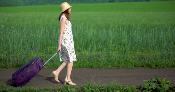 A Girl in a Dress and a Hat Walks Through a Green Field of Grass alt