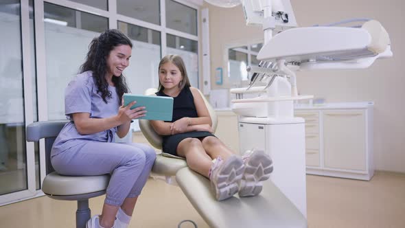 Wide Shot Cheerful Young Female Dentist and Adolescent Patient Talking Using Tablet Smiling in Slow alt