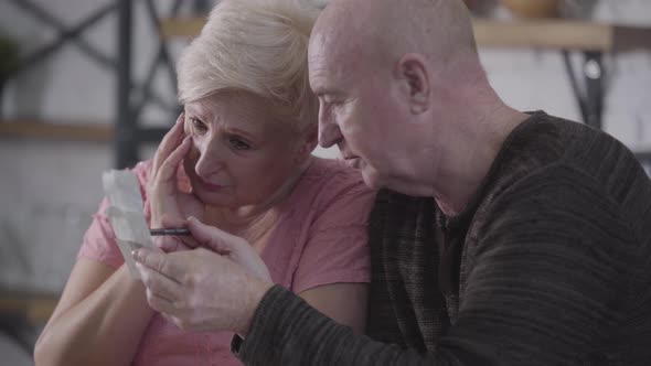 Close-up of Old Caucasian Man Explaining Overspending To His Wife. Upset Woman Holding Head with alt