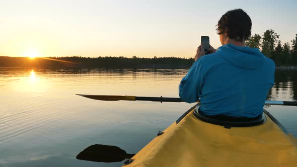 Man Takes a Photo of Sunset on Smartphone on Lake on Kayak with Paddle, Self-isolation Outdoor alt