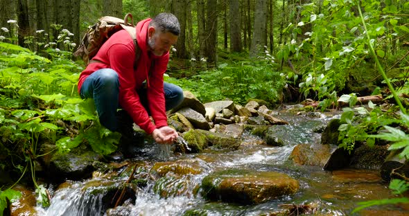 A Bearded Man with a Backpack Drinks Water From a Mountain Stream alt