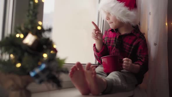 Cute Caucasian Boy in Santa Had and Flannel Shirt Sitting on Window Sill Drinking Milk From Cup