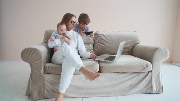 Pleasant Young Mother Freelancer Working on Computer Sitting on a Sofa with Cute Baby Infant at Home alt