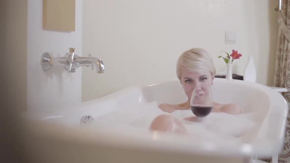Relaxed Blond Woman Drinking Red Wine in Bathtub with Bath Foam. Portrait of Happy Caucasian Lady alt