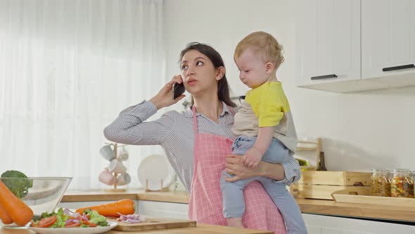Caucasian busy mother doing housework with baby boy toddler in kitchen. alt