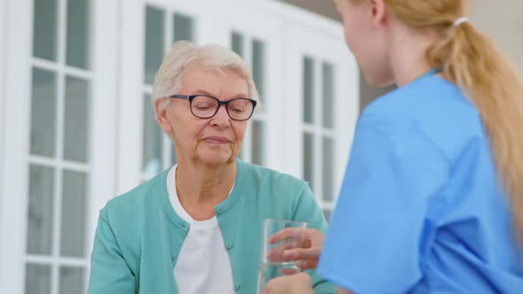 Careful nurse with ponytail talks to senior woman resident and gives glass alt