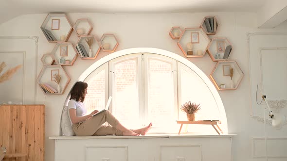 a Young Woman Sits on the Windowsill in a Cozy Bright Apartment and Communicates Via Video alt