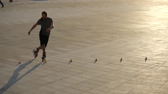 Man Roller Skater with Long Hair Dancing Between Cones Makes Rotations in Evening City at Sunset alt