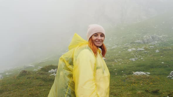 Medium shot of woman in raincoat turns to the camera then continues her way up in the mountains. alt