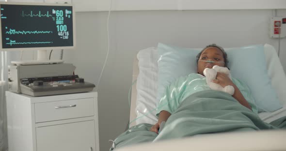Afroamerican Kid Lying in Hospital Bed with Oxygen Tube and Plush Toy alt