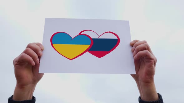 Man Hands Holding of Heart Drawingon and Russian Flag and Ukraine Flag the Cardboard alt