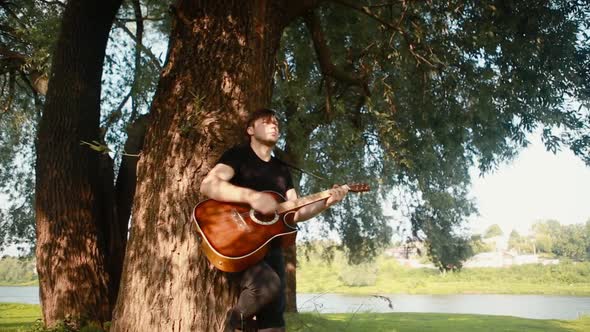 A guy stands near a tree and plays the guitar.