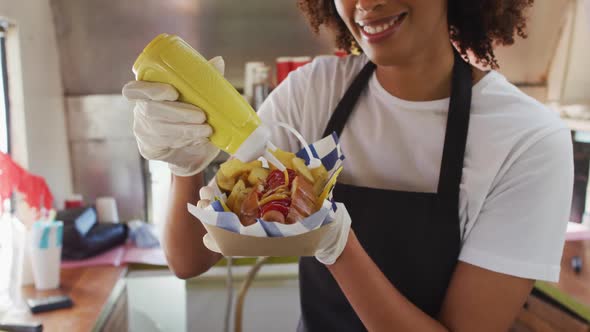 African american woman wearing apron putting mustard over the hot dog in food truck alt