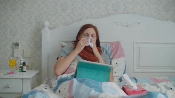 Sick Young Woman Using Tablet Computer Laying in Bed. Woman with Warm Scarf on Neck Coughs and Blows alt