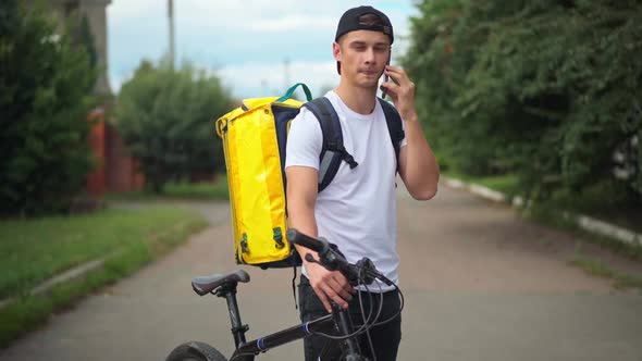 Caucasian Courier Talking on Phone with Client Standing on Suburban Road with Bike and Yellow alt