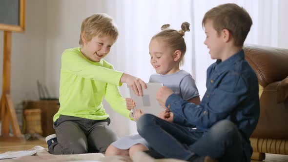 Three Cheerful Children Fighting for Tablet Sitting in Living Room at Home alt