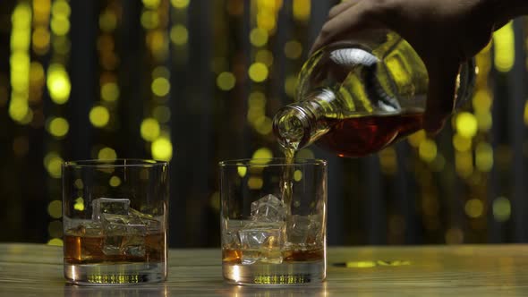 Pouring of Whiskey, Cognac or Brandy From Bottle Into Glasses with Ice Cubes. Shiny Background alt