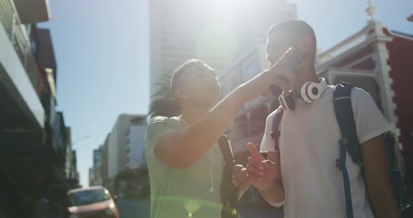 Two happy mixed race male friends standing, talking and using smartphone in the street alt
