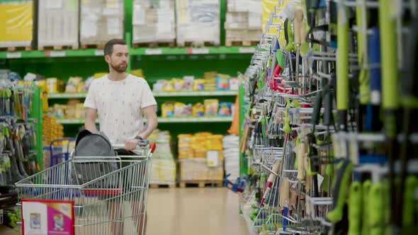 Alone Young Man is Walking in Sales Hall in Shop Rolling Trolley in Front alt