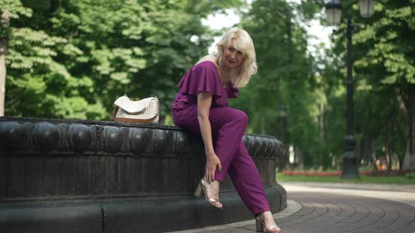 Wide Shot Adult Tired Woman Rubbing Ankle Sitting at Fountain in Summer Park alt